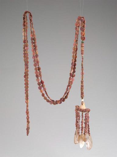 An ornate, handcrafted necklace is suspended against a neutral grey background. The necklace is made of long strands of small, flat, disc-shaped beads in varying shades of pink and red ochre. It hangs from two points, creating a draped loop. The strand on the left hangs straight down. The strand on the right is shorter and features a tassel-like pendant. This pendant consists of a thicker, beaded section leading down to a light-colored, cone-shaped element. Dangling from the base of the cone are several shorter strings of the same reddish beads, each ending with a larger, translucent, shell-like pendant [gen AI description].