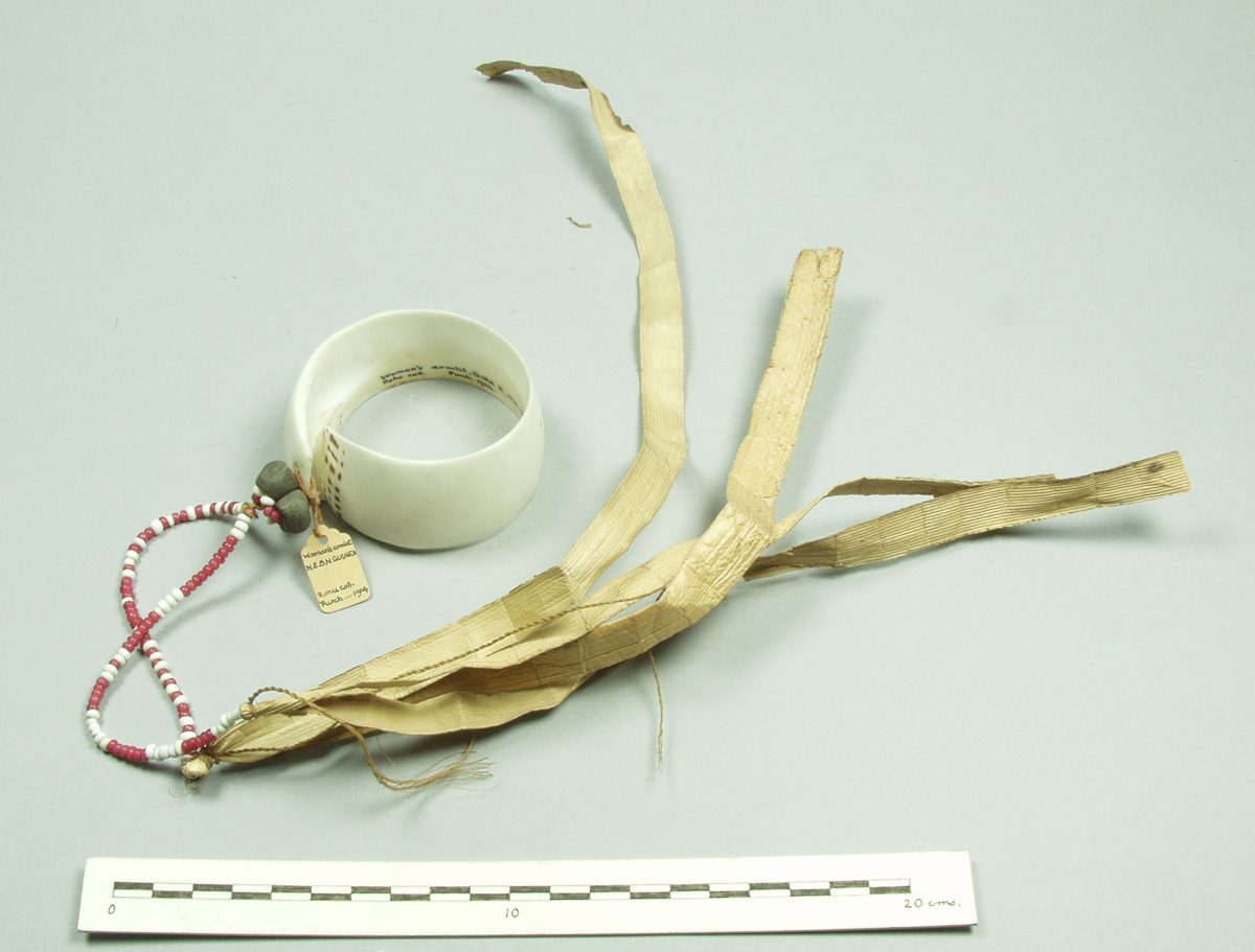 Arm ornament; Cylindrical-spiral of conus shell, with 2 perforations through which is tied 2 plant fibre cords strung with red & white glass beads, 3 black banana seed pods and long strips of palm leaf. 1904. Pitt Rivers Museum, University of Oxford, 1904.60.131. https://www.prm.ox.ac.uk/collections-online#/item/prm-object-99833.