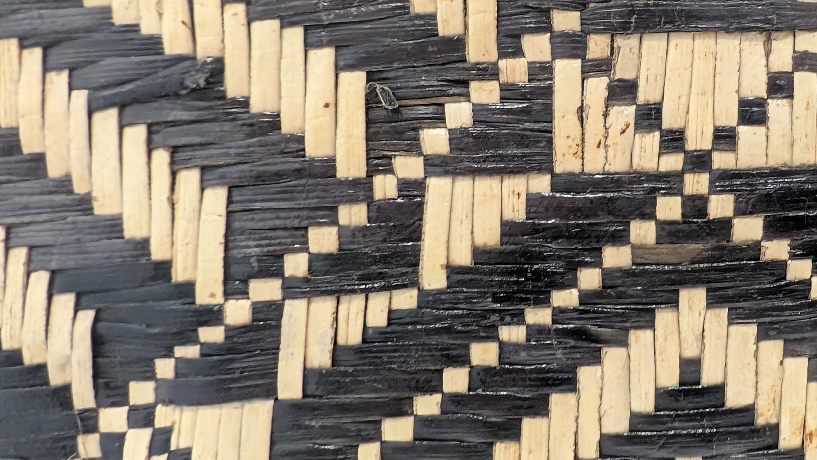 An extreme close-up photograph of a basket woven from black-lacquered and natural beige colored strands of dried pandanus in a pattern of intersecting diagonal stripes.