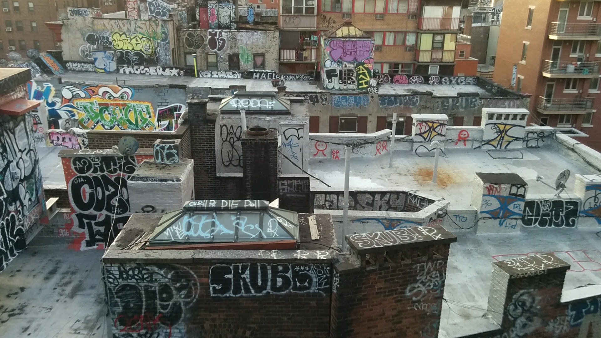 A high-angle daytime view shows a dense cluster of urban rooftops heavily covered in many layers of colorful and intricate graffiti, with brick buildings of various heights filling the frame, creating a gritty, layered, and expressive urban environment [gen AI description].